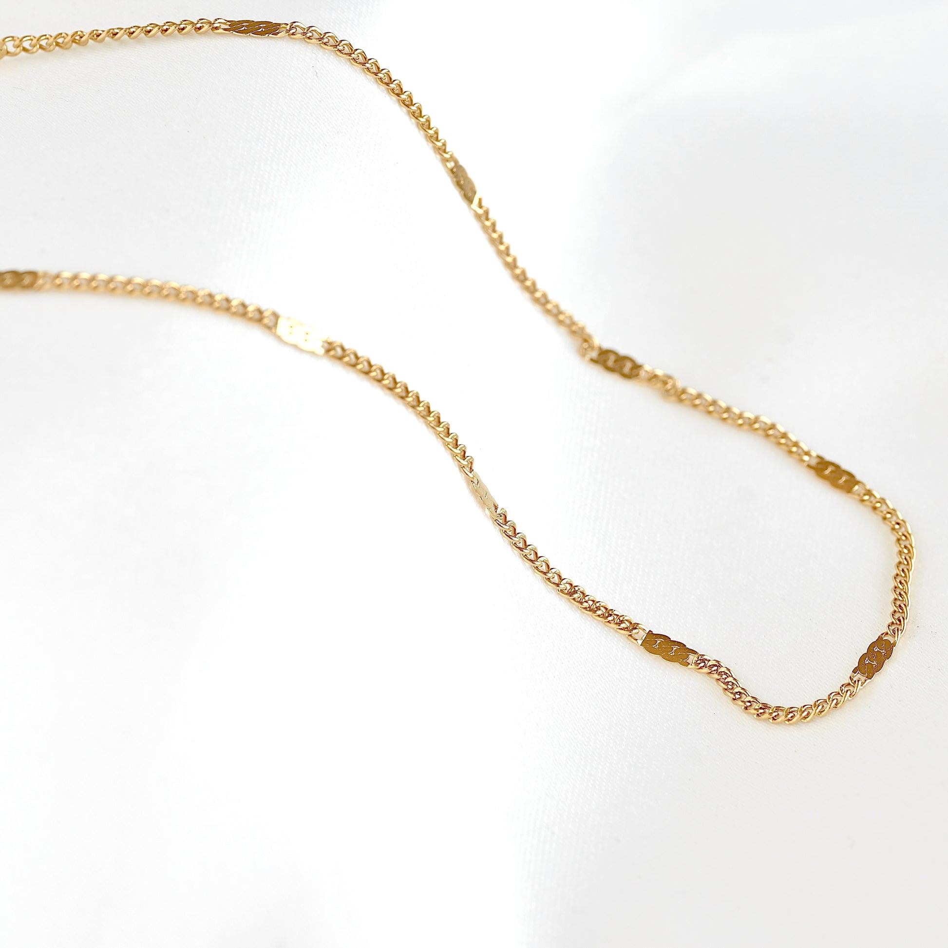 A delicate gold chain necklace with small rectangular links, displayed on a soft white fabric background.