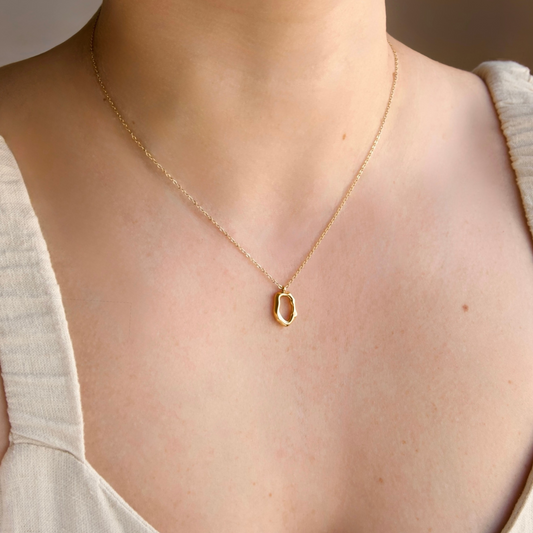 A delicate gold chain necklace with an abstract, open oval pendant, worn on a model's neck over a light beige top.
