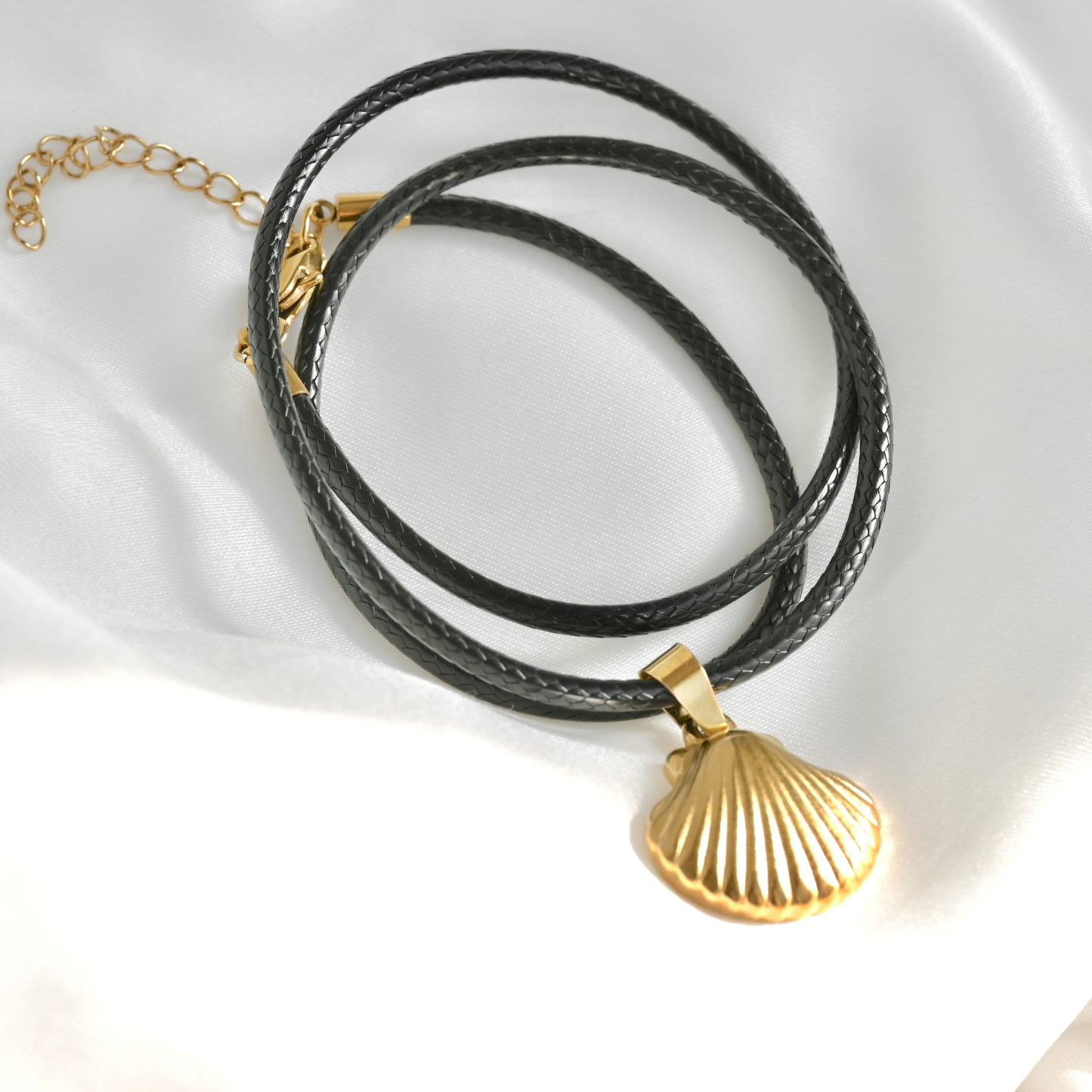 A black braided cord necklace with a gold-toned shell-shaped pendant, displayed on a white satin fabric background.