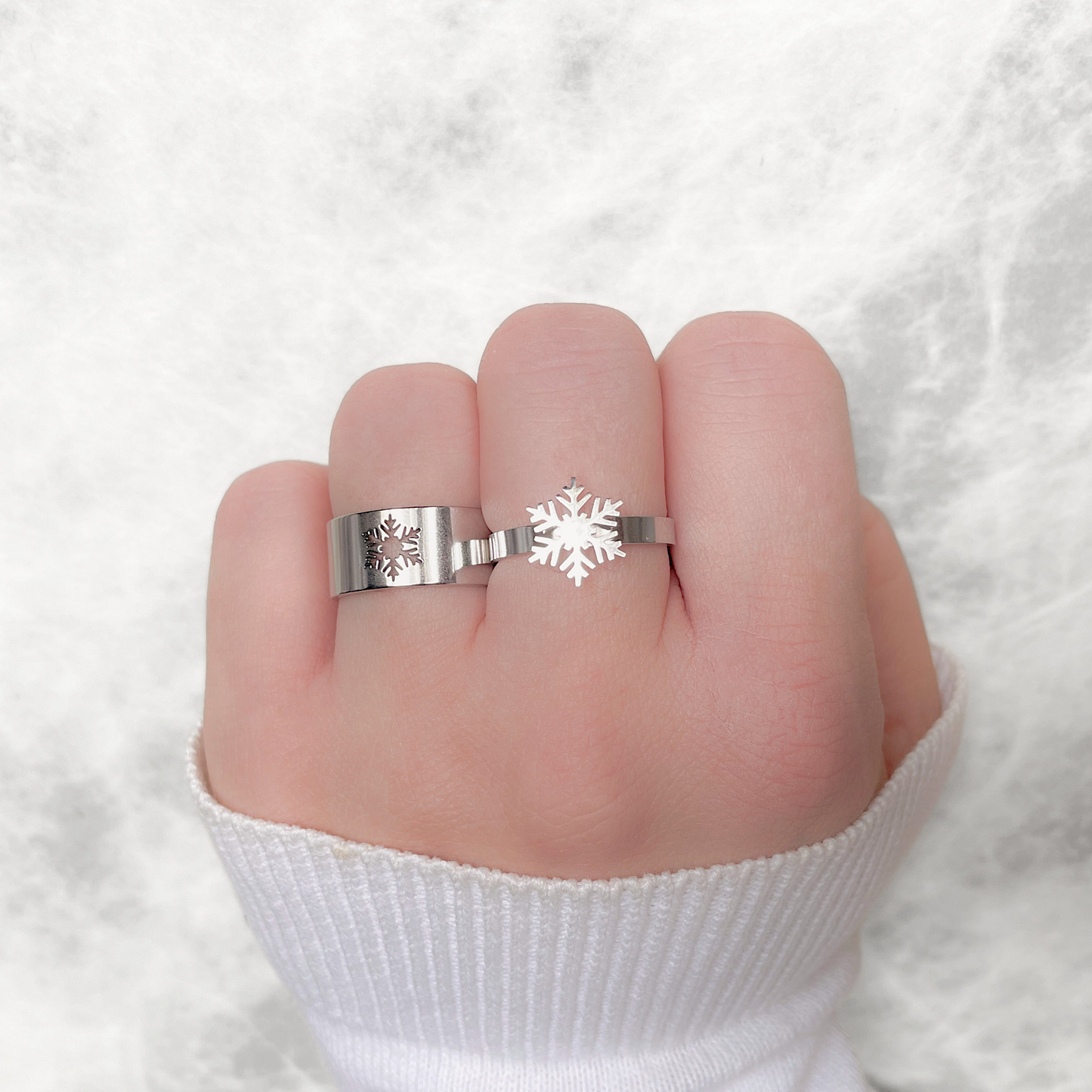 Top wide angled view of silver ring with snowflake charm and wide flat band with cutout snowflake design on a finger against a white background