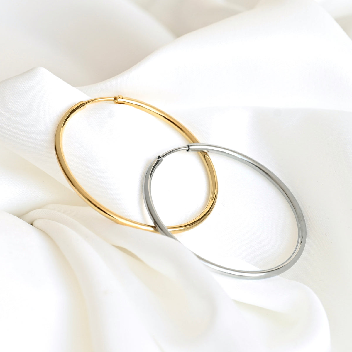 Two large hoop earrings, one in polished gold and one in polished silver, interlocking on a draped white fabric background.