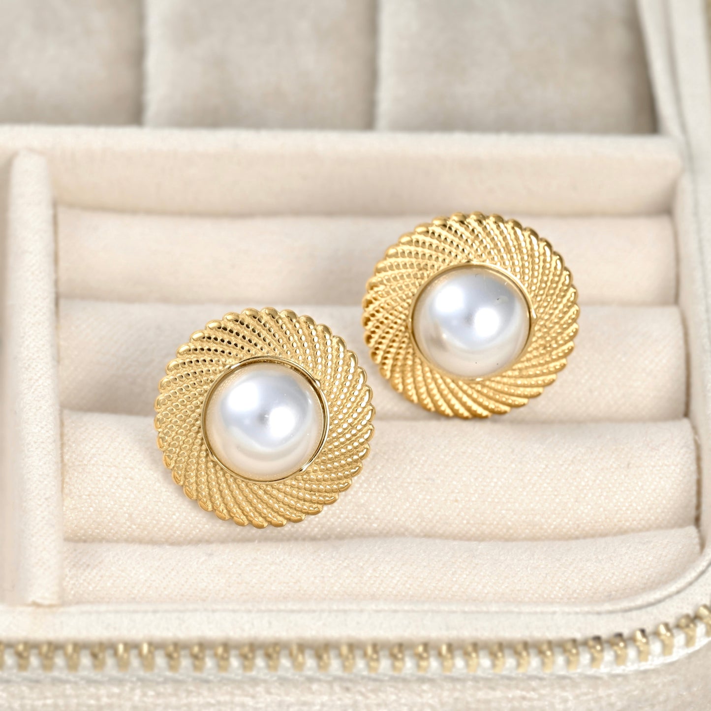 A pair of round gold-tone earrings with textured spiral edges and white pearl centers, displayed in a cream-colored jewelry box.