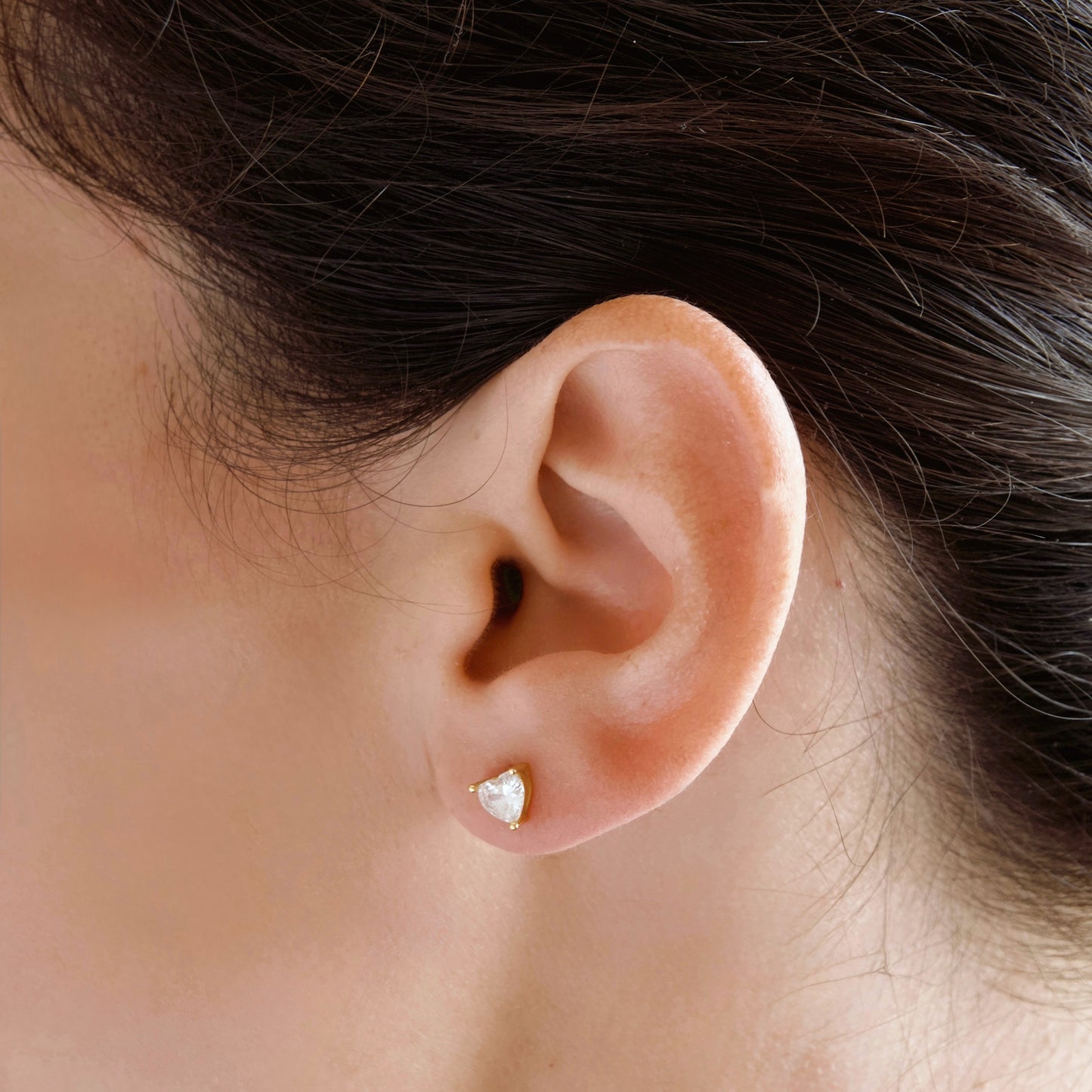 A close-up of a person's ear wearing a small gold stud earring with a heart-shaped clear cubic zirconia stone.