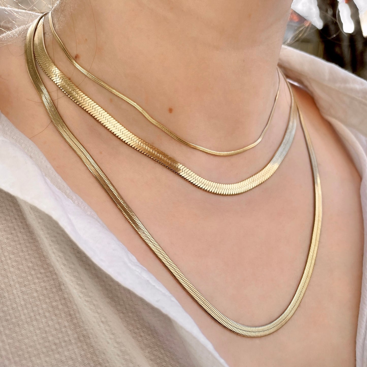 Three gold herringbone chain necklaces with 2mm, 6mm and 4mm widths with various lengths layered on a person with a neutral background