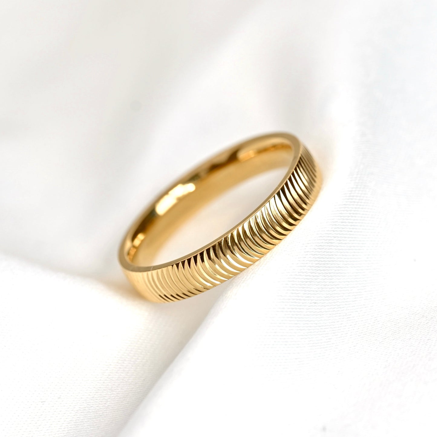 A gold ring with a textured, ridged outer band, resting on a soft, white draped fabric background.
