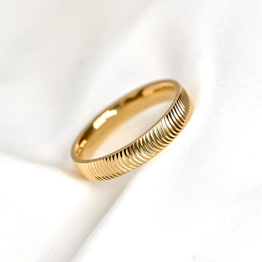 A gold ring with a textured, ridged outer band, resting on a soft, white draped fabric background.