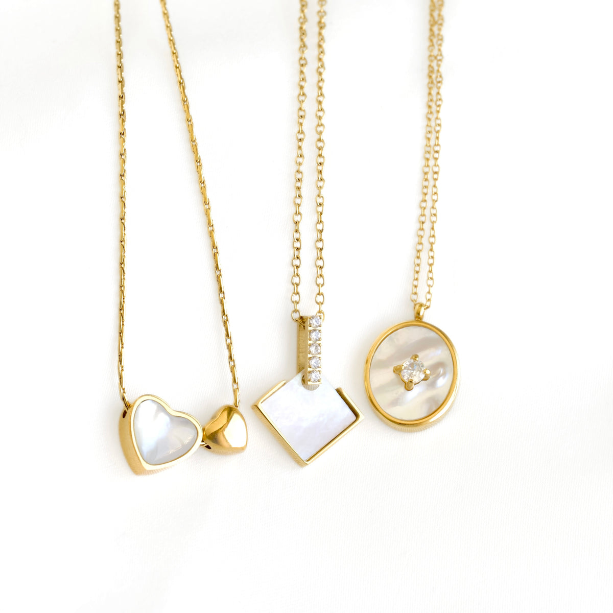 Three gold chain necklaces with mother-of-pearl pendant charms in heart, square, and oval shapes, displayed on a clean white background.