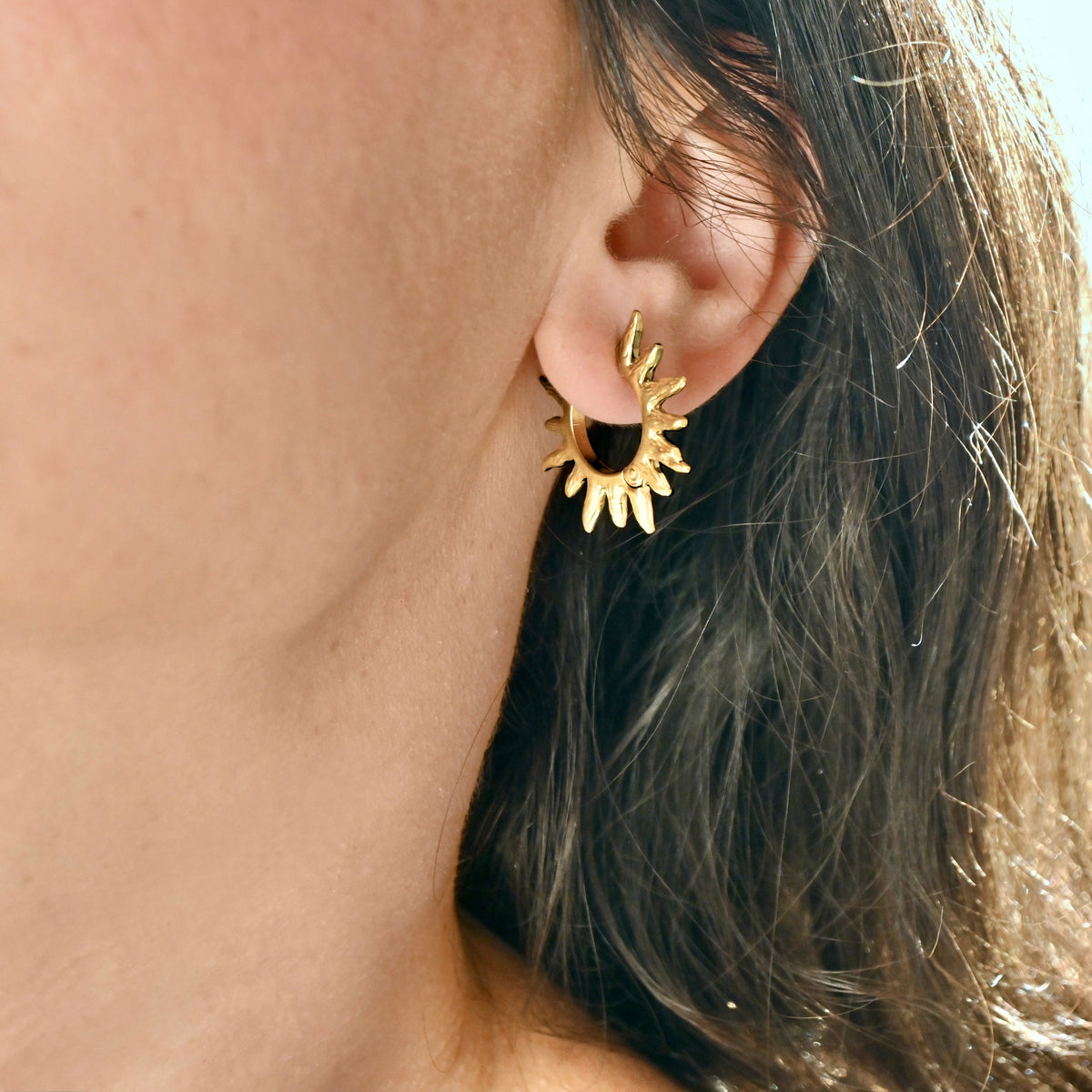 A close-up of a person's ear wearing a medium gold sun hoop earring with textured petal-like details.