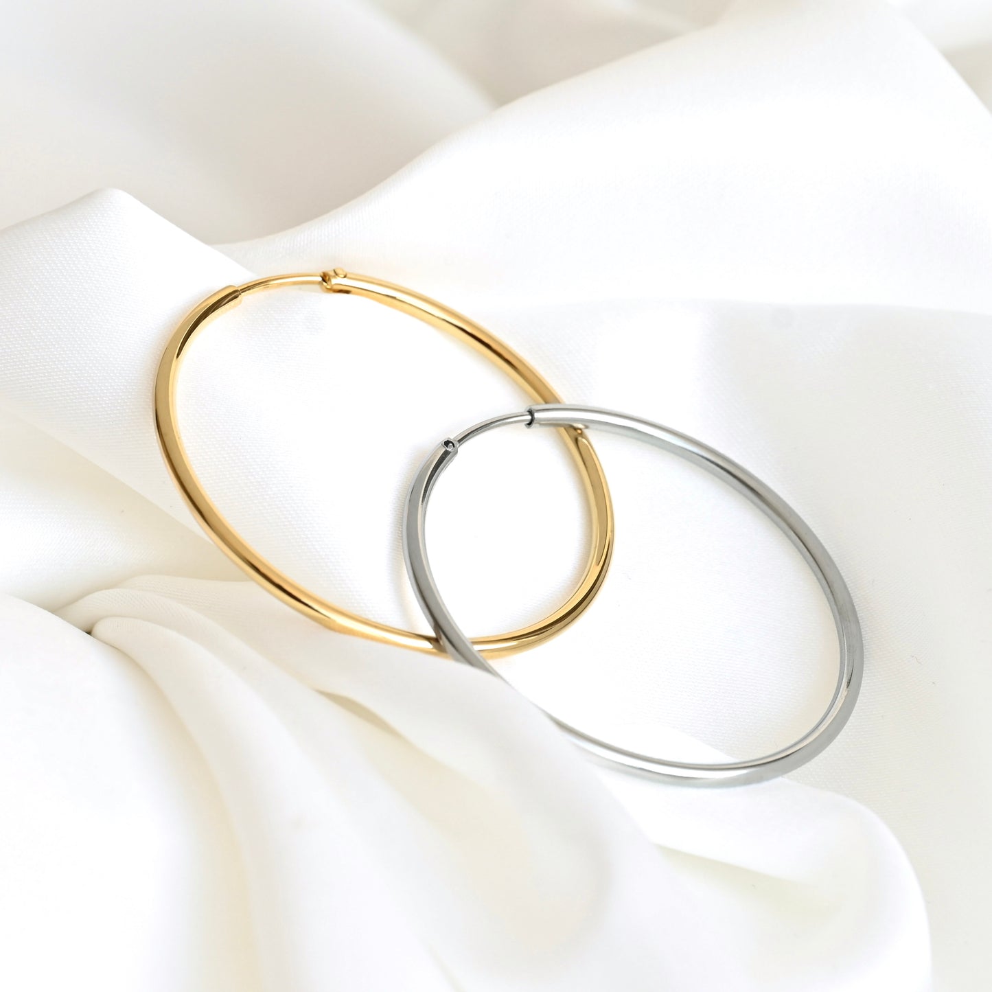 Two large hoop earrings, one in polished gold and one in polished silver, interlocking on a draped white fabric background.