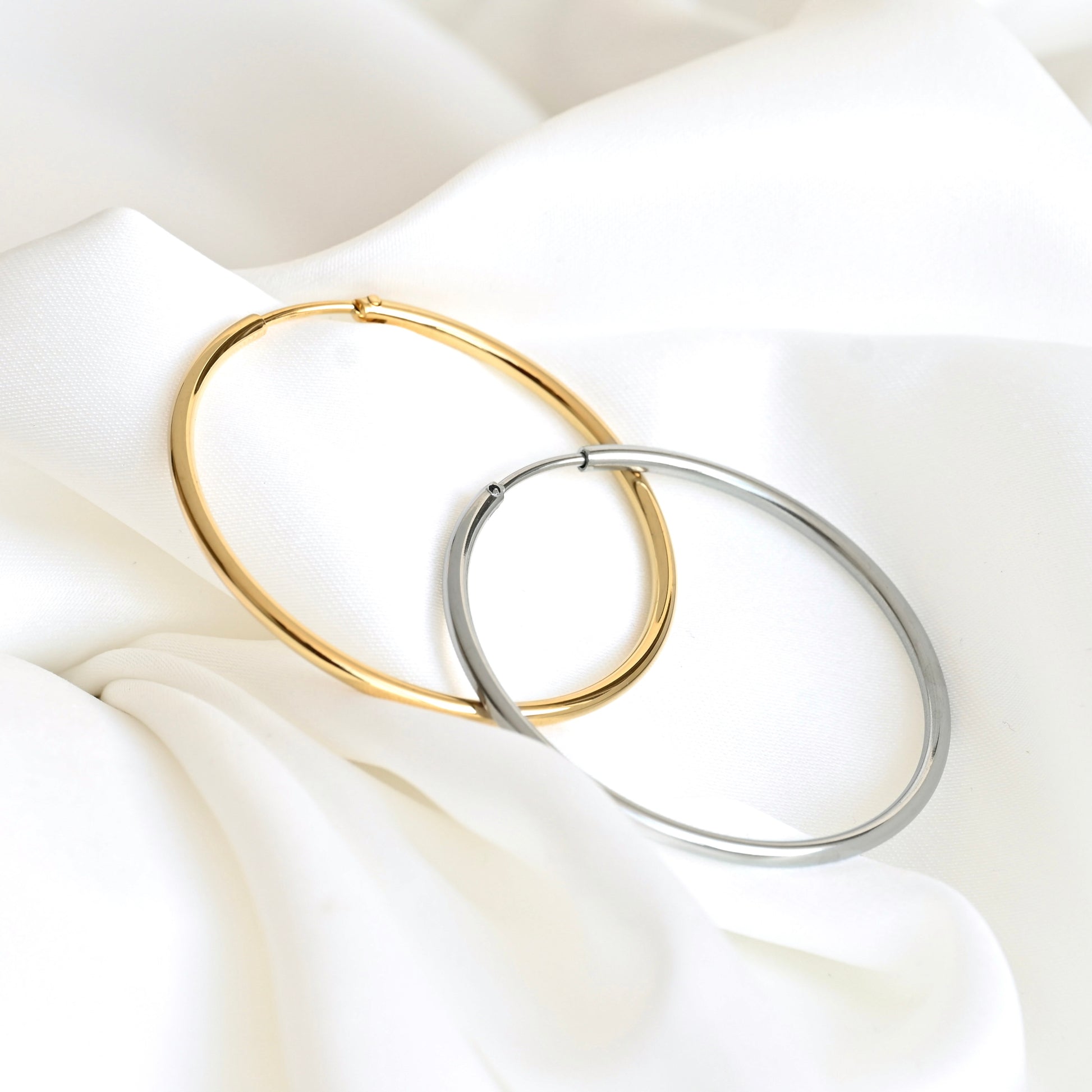 Two large hoop earrings, one in polished gold and one in polished silver, interlocking on a draped white fabric background.
