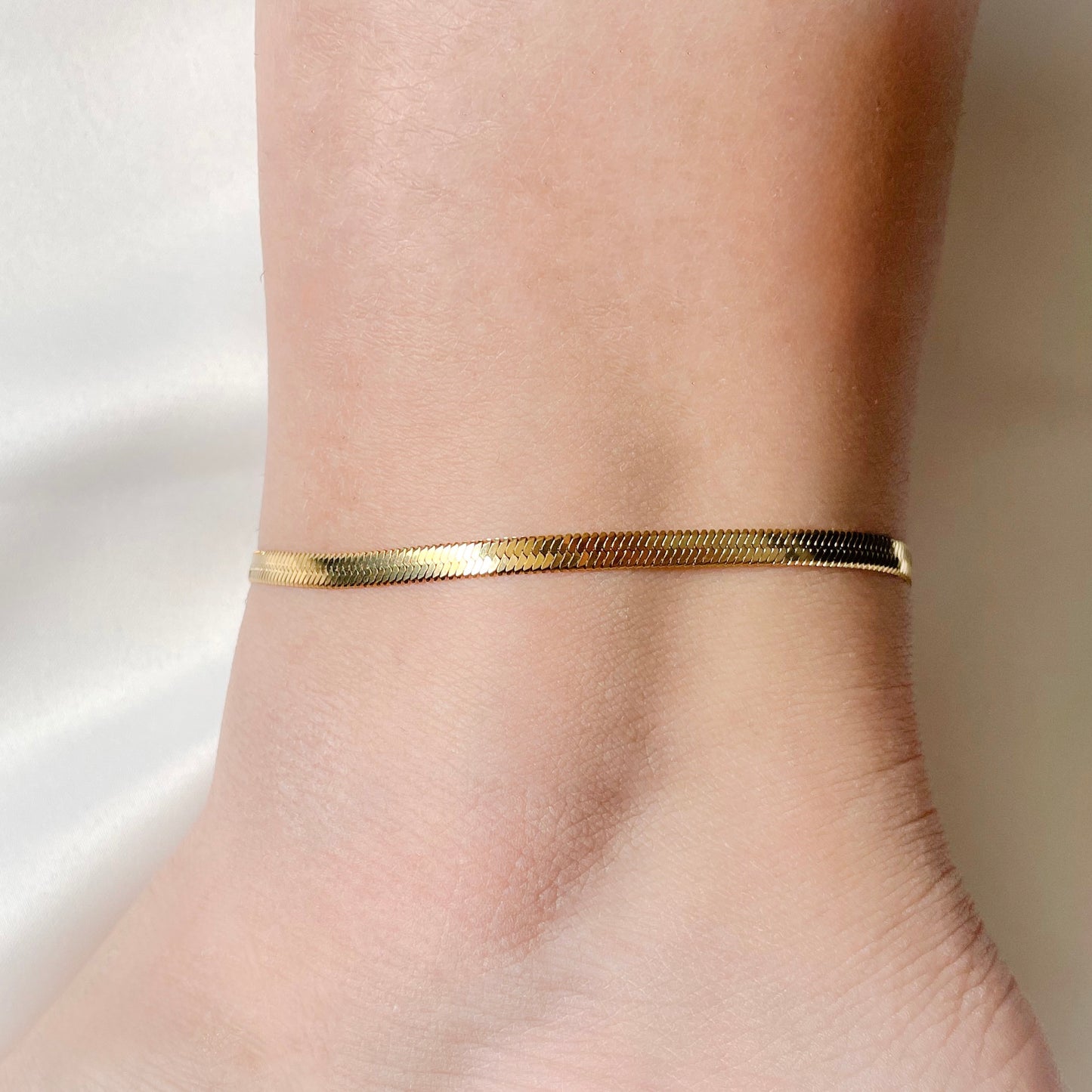 Gold 4mm herringbone anklet on a person's ankle against a neutral background