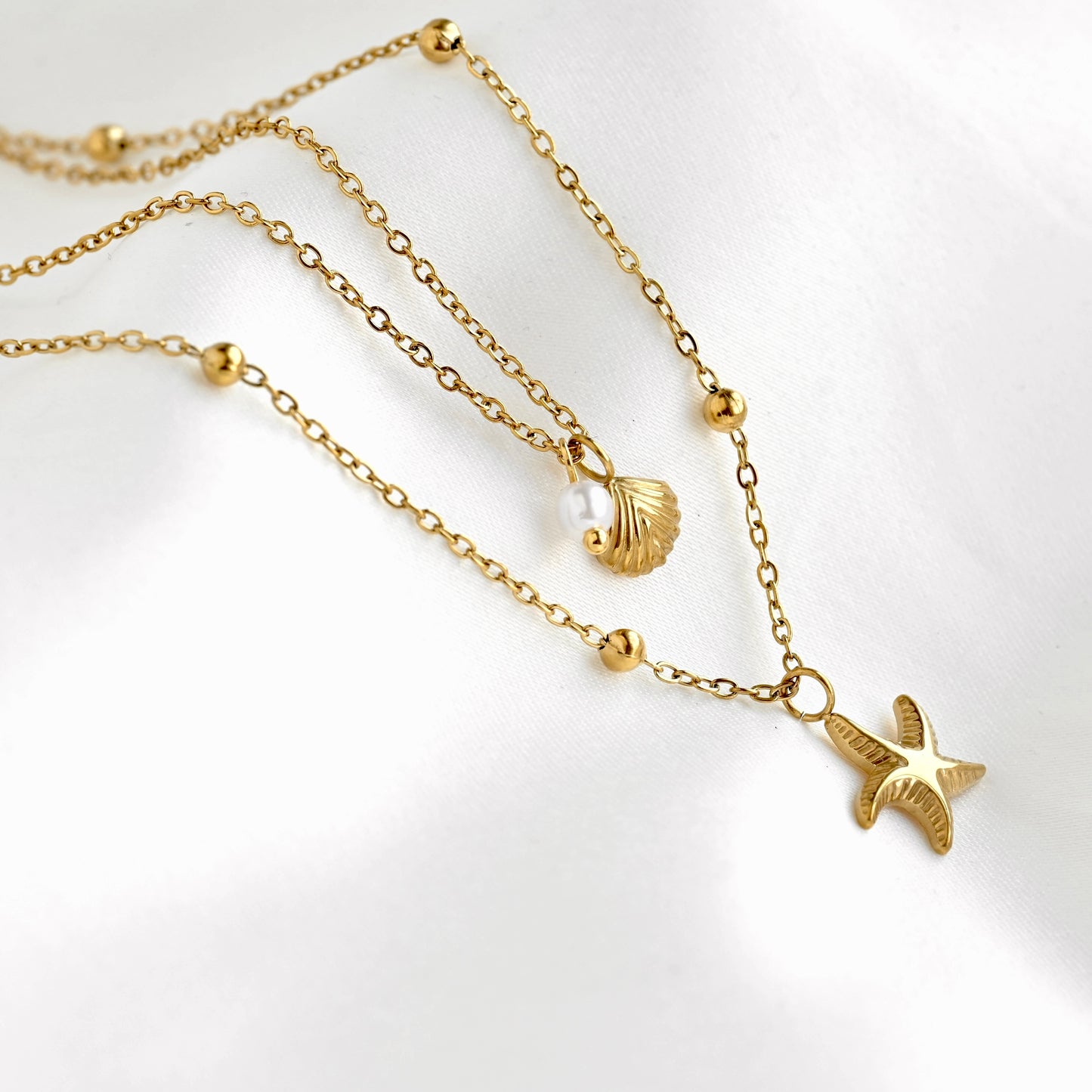 Side viw of a gold layered necklace featuring a starfish pendant on a satellite chain and a seashell charm & pearl on a cable chain on a white background.