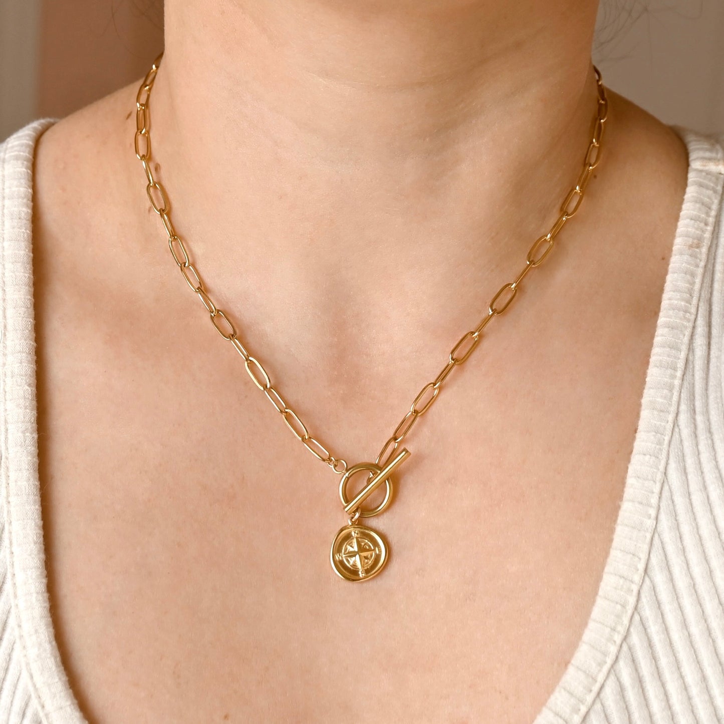 A gold chain necklace with a toggle clasp and a circular pendant featuring a double-G style emblem, shown worn on a model's neck over a white ribbed top.