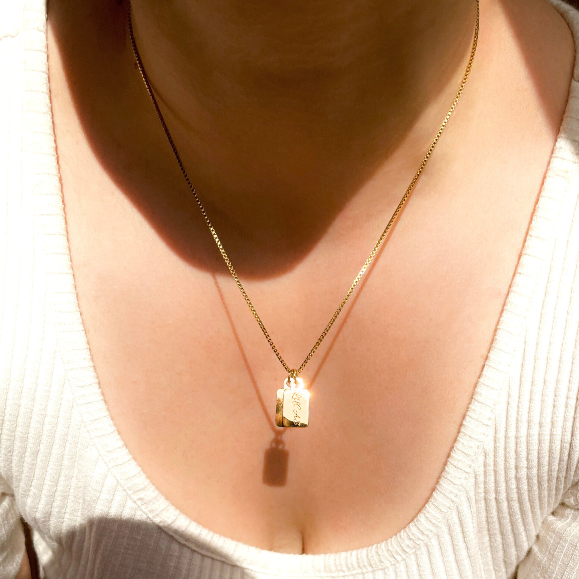 A close-up of a person's neck and chest wearing a delicate gold chain necklace with two small rectangular pendants engraved with names, styled with a white ribbed knit top.