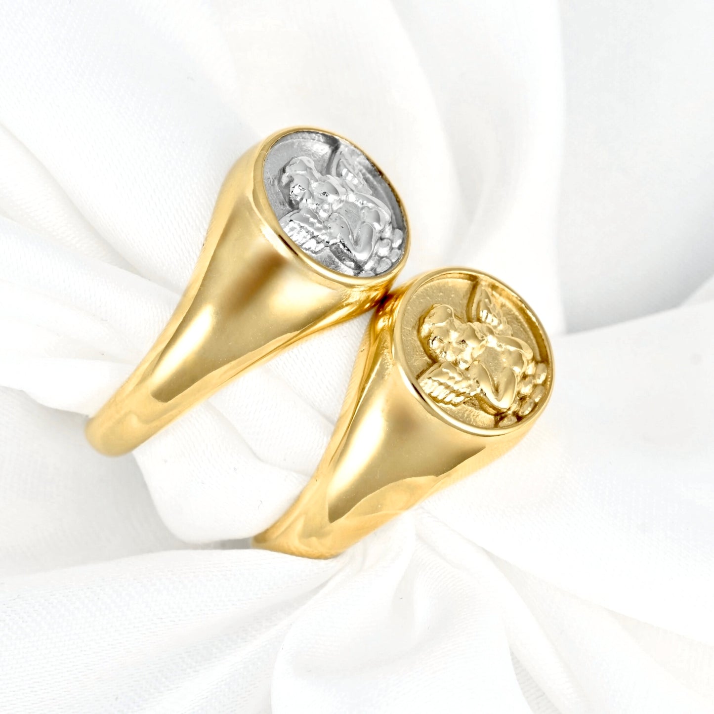 Two gold signet rings with round cabochons, one featuring a silver-toned angel motif, displayed on a white fabric background.