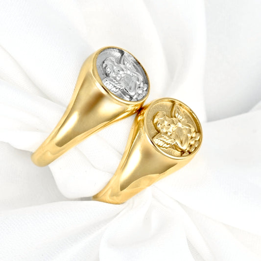 Two gold signet rings with round cabochons, one featuring a silver-toned angel motif, displayed on a white fabric background.