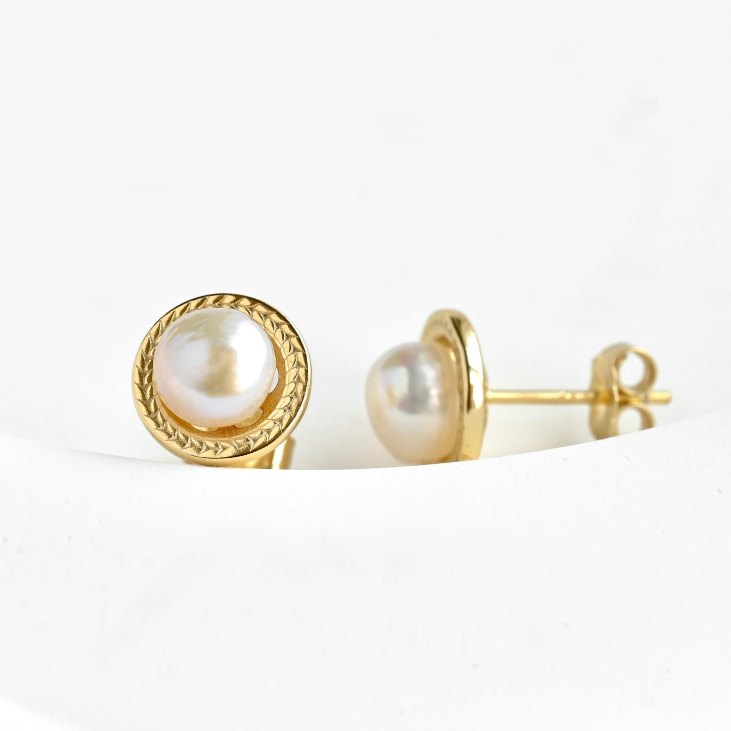 A pair of gold stud earrings with round white freshwater pearls, one shown from the front and the other from the side/back, resting on a white curved surface.