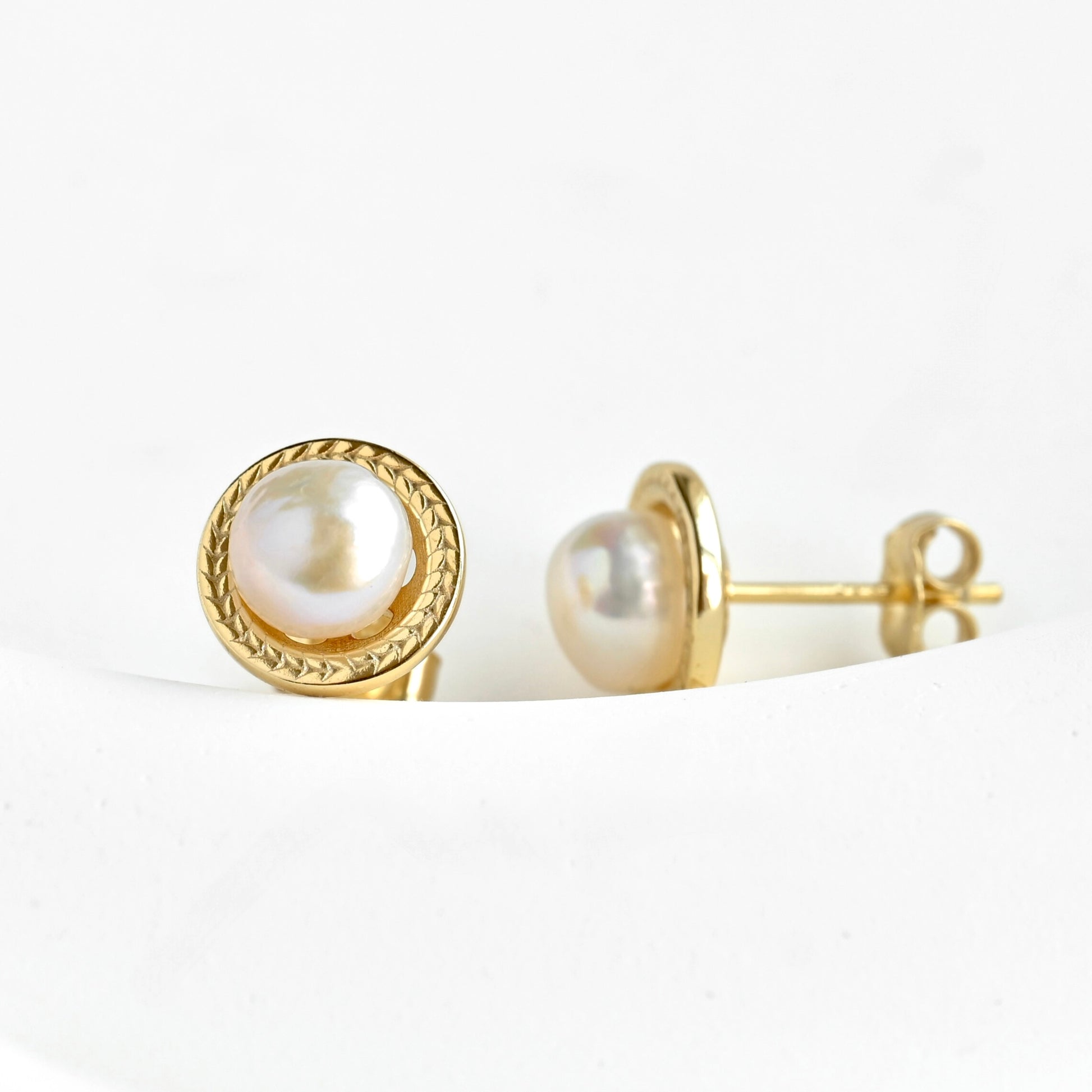 A pair of gold stud earrings with round white freshwater pearls, one shown from the front and the other from the side/back, resting on a white curved surface.