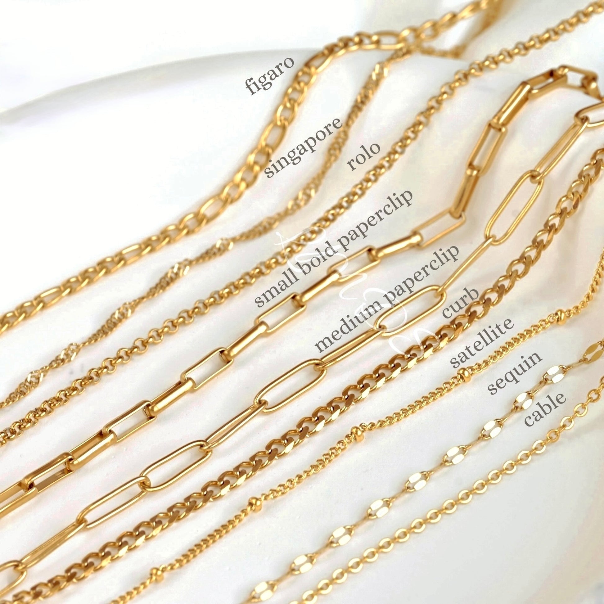 Gold chains with various designs on a white background. The chain types are as follows from back to front: figaro, singapore, rolo, small bold paperclip, medium paperclip, curb, satellite, mariner and cable.