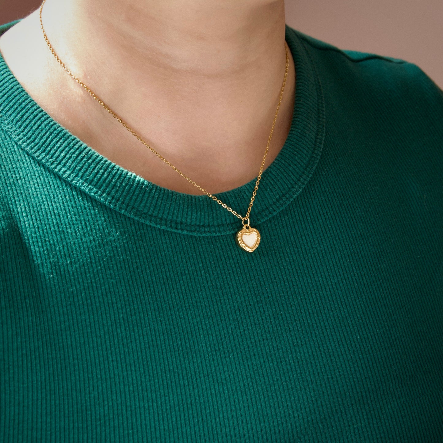 A close-up of a person wearing a delicate gold chain necklace with a small heart-shaped pendant, paired with a dark green ribbed knit top.