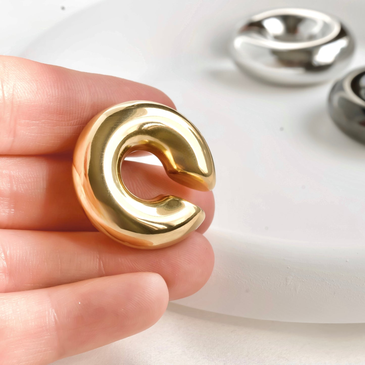 Gold dome ear cuff held between fingers with silver earrings in the background.