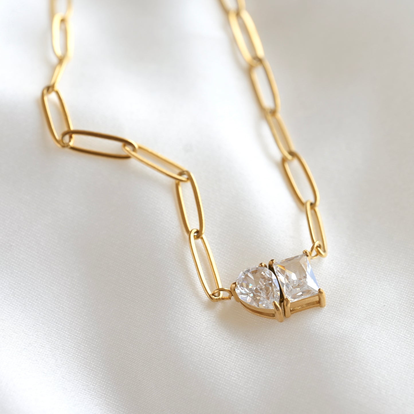 A gold paperclip chain necklace with a pendant featuring two clear teardrop and rectangle CZ stones, displayed on a soft white fabric background.