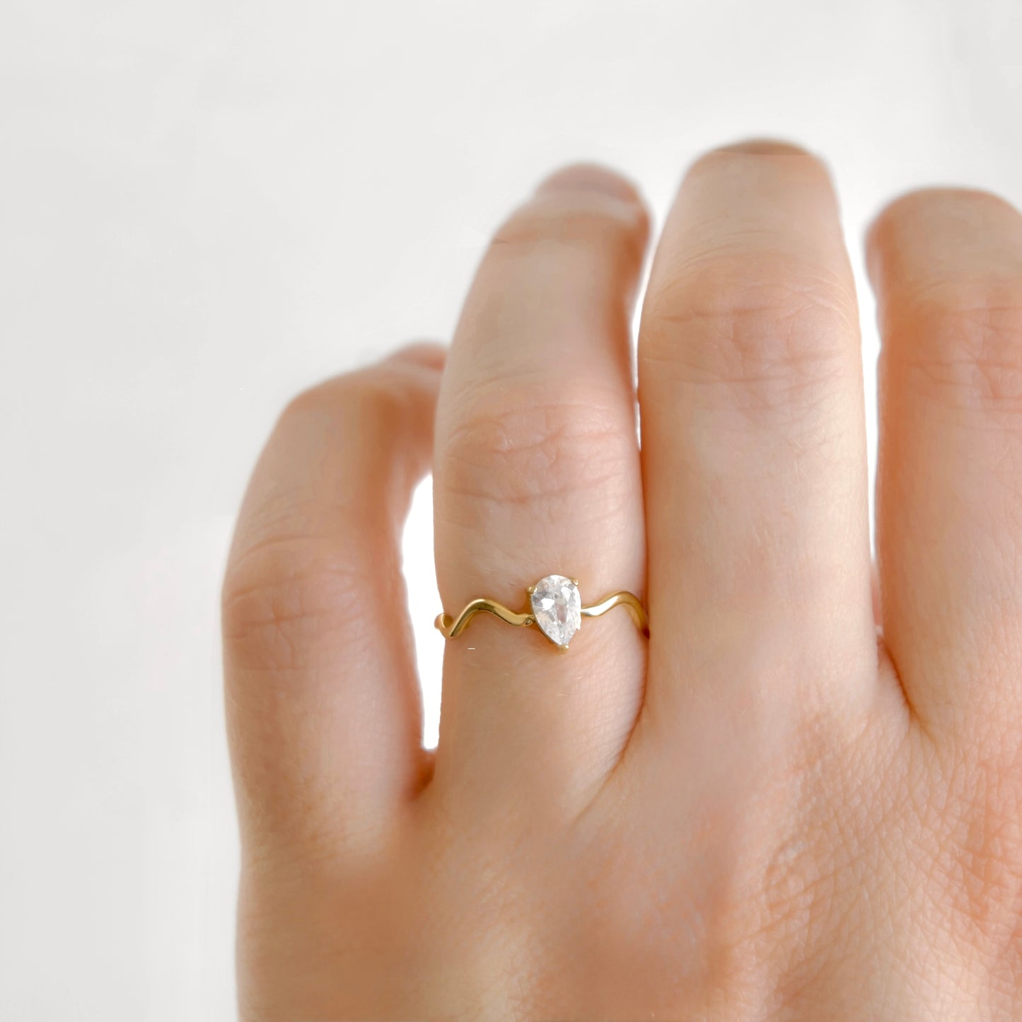 A close-up of a hand wearing a delicate yellow gold ring with a small pear-cut clear gemstone, set against a soft, light off-white background.