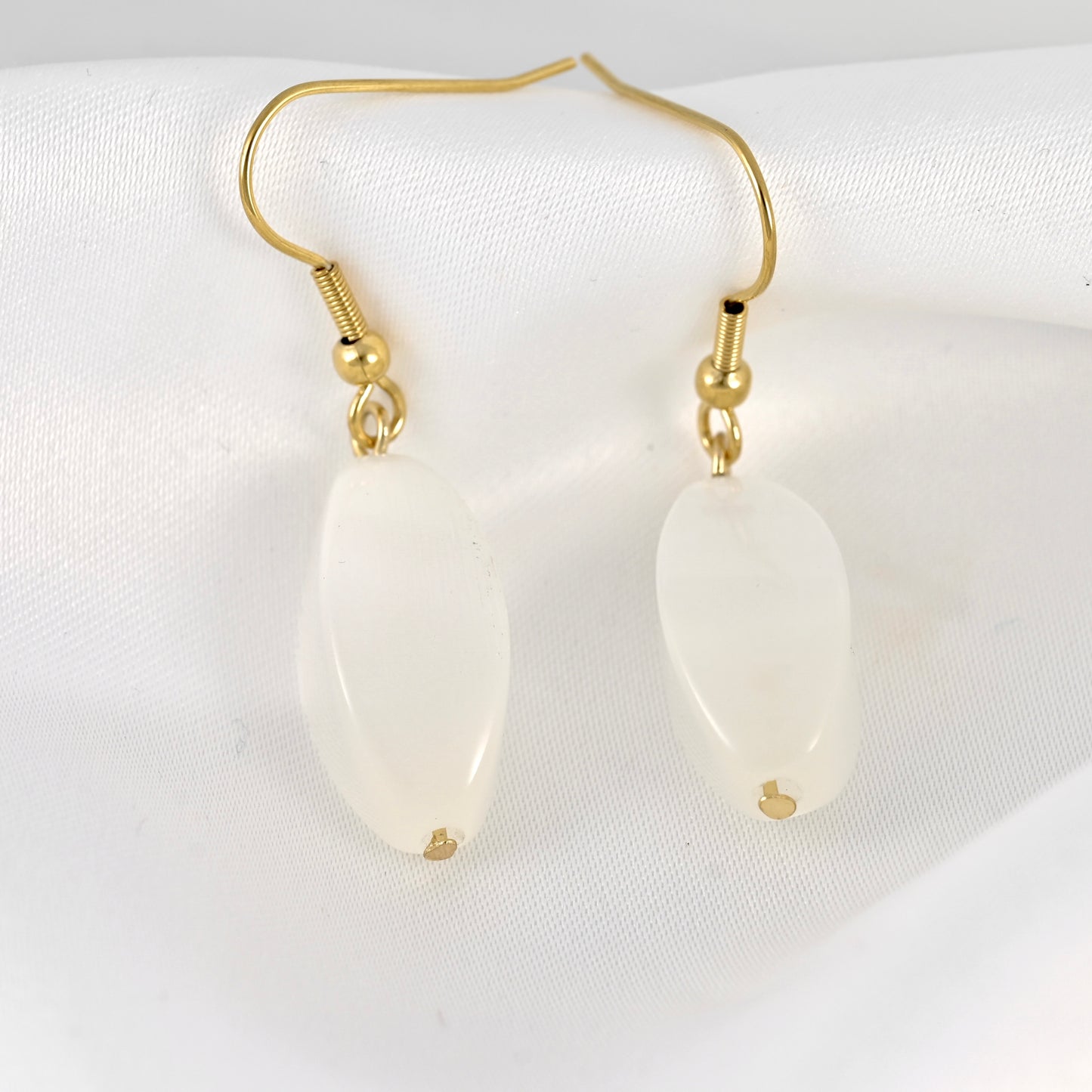 A pair of gold drop earrings with smooth, teardrop-shaped white stones, displayed on a textured white fabric background.