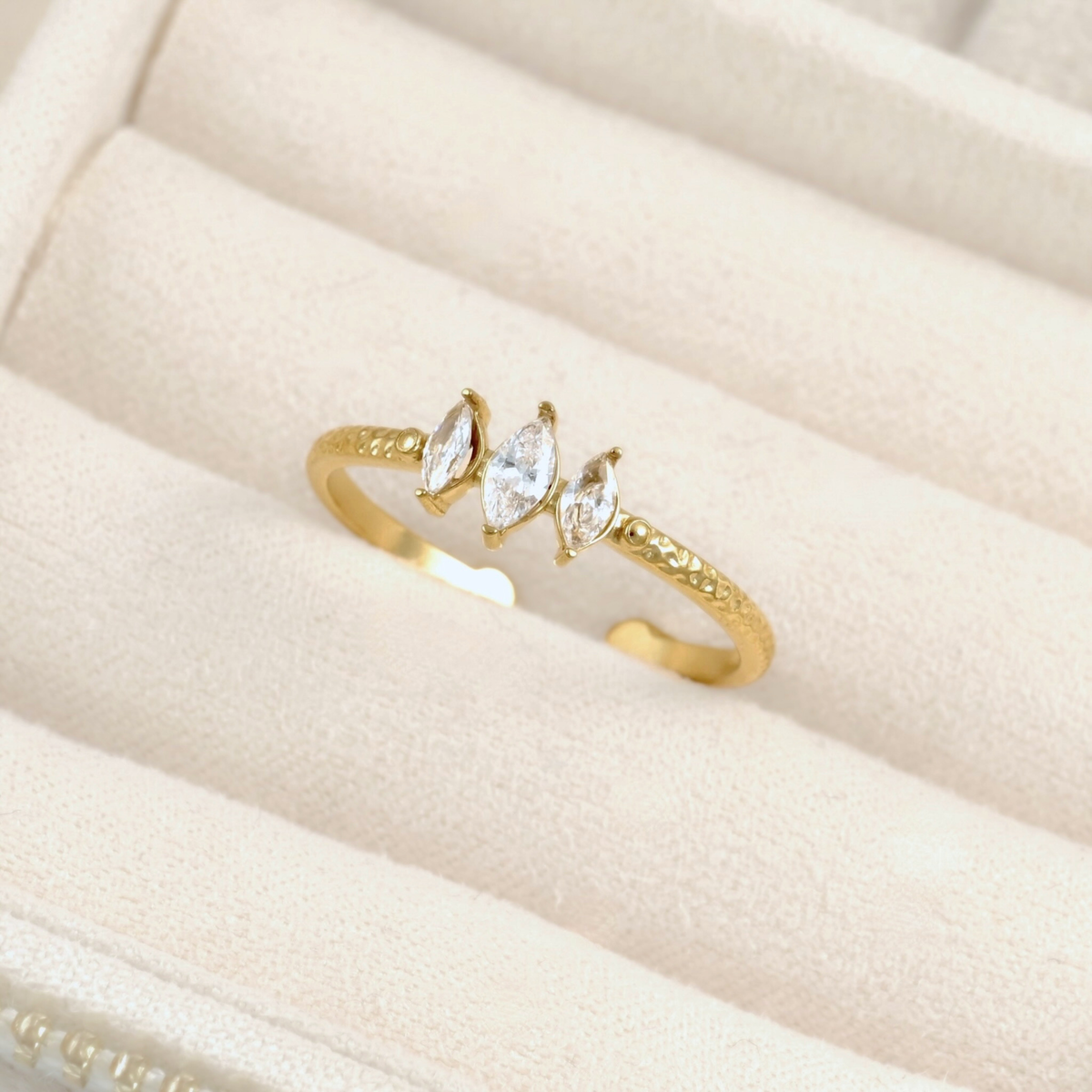 A gold open-bend ring with three marquise-cut clear gemstones, displayed on a cream-colored jewelry box cushion.