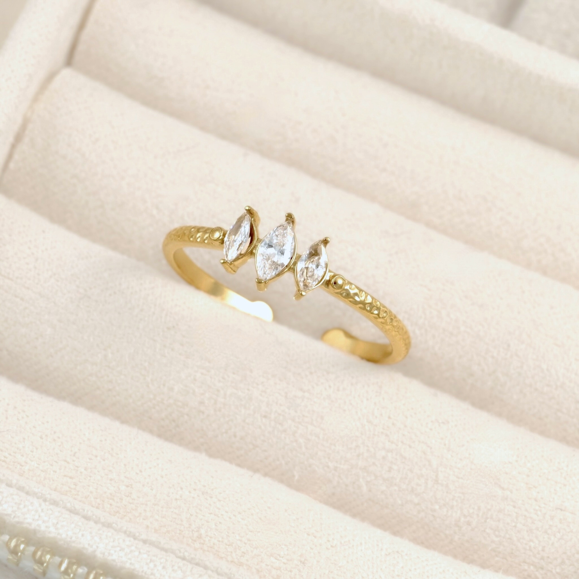 A gold open-bend ring with three marquise-cut clear gemstones, displayed on a cream-colored jewelry box cushion.