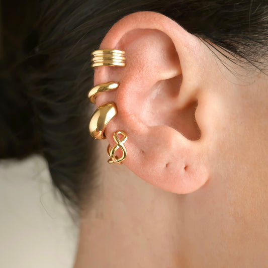 Close-up of an ear wearing gold ear cuff stack including oval links, chunky dome, dainty dome and chunky striped designs with a blurred background