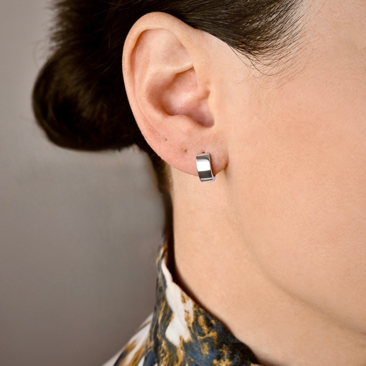 A close-up of a person's ear wearing  small thick polished silver huggie hoop earrings.