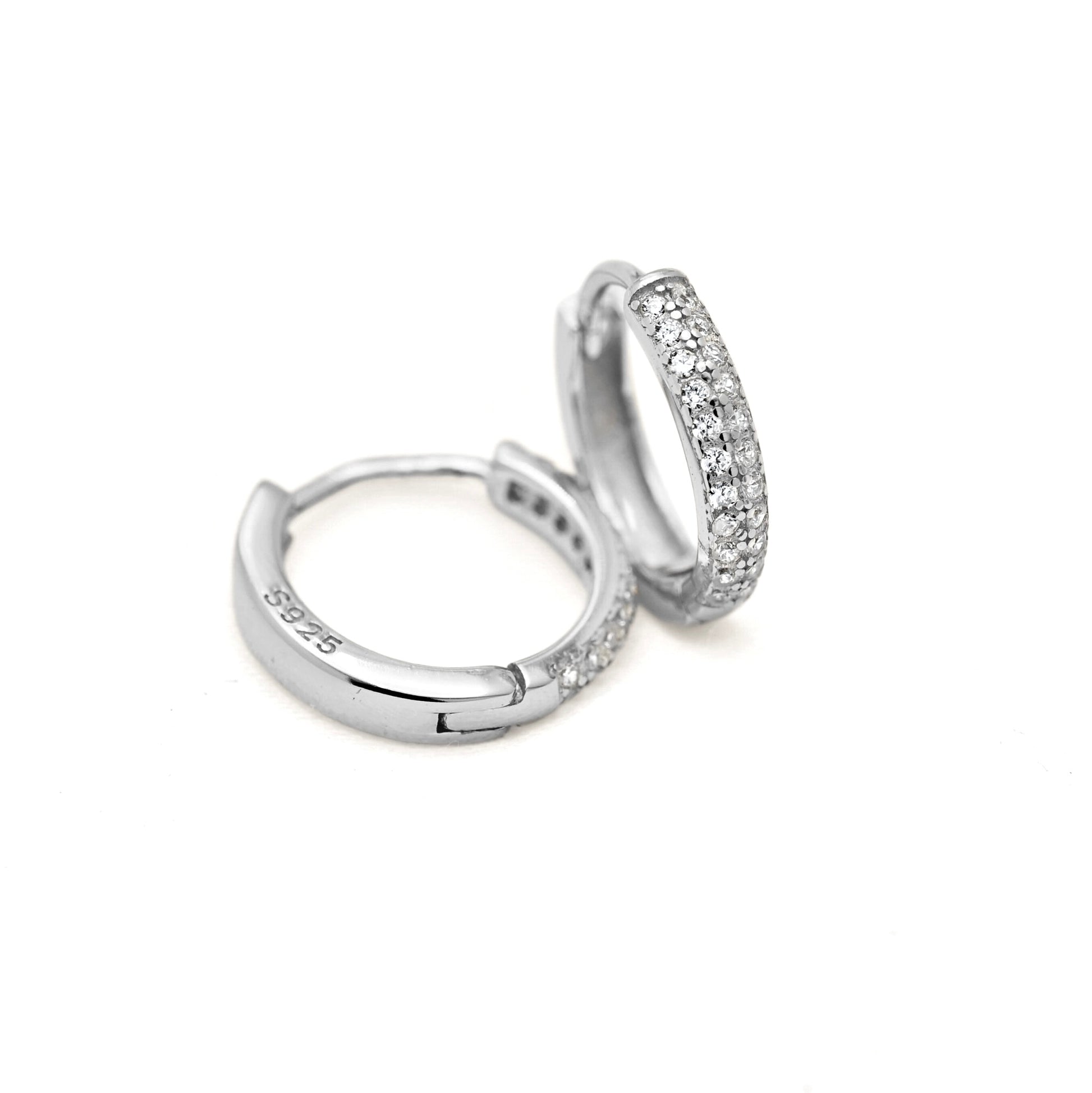 A pair of silver huggie hoop earrings, one showing the plain polished interior and the other set with a row of clear pavé stones along the front, on a white background.
