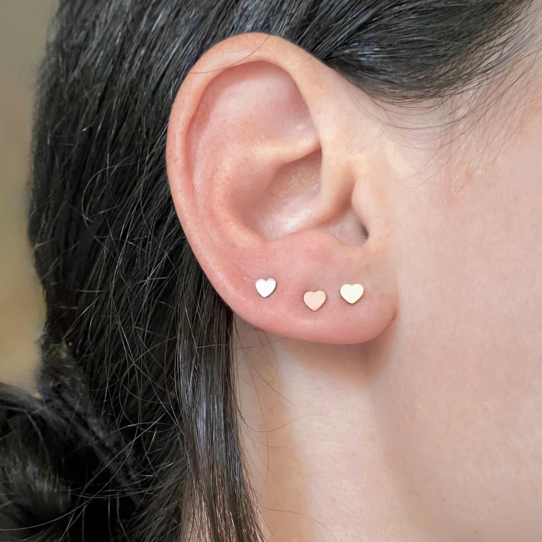 Close-up of an ear wearing three heart-shaped earring in gold, rose gold and silver