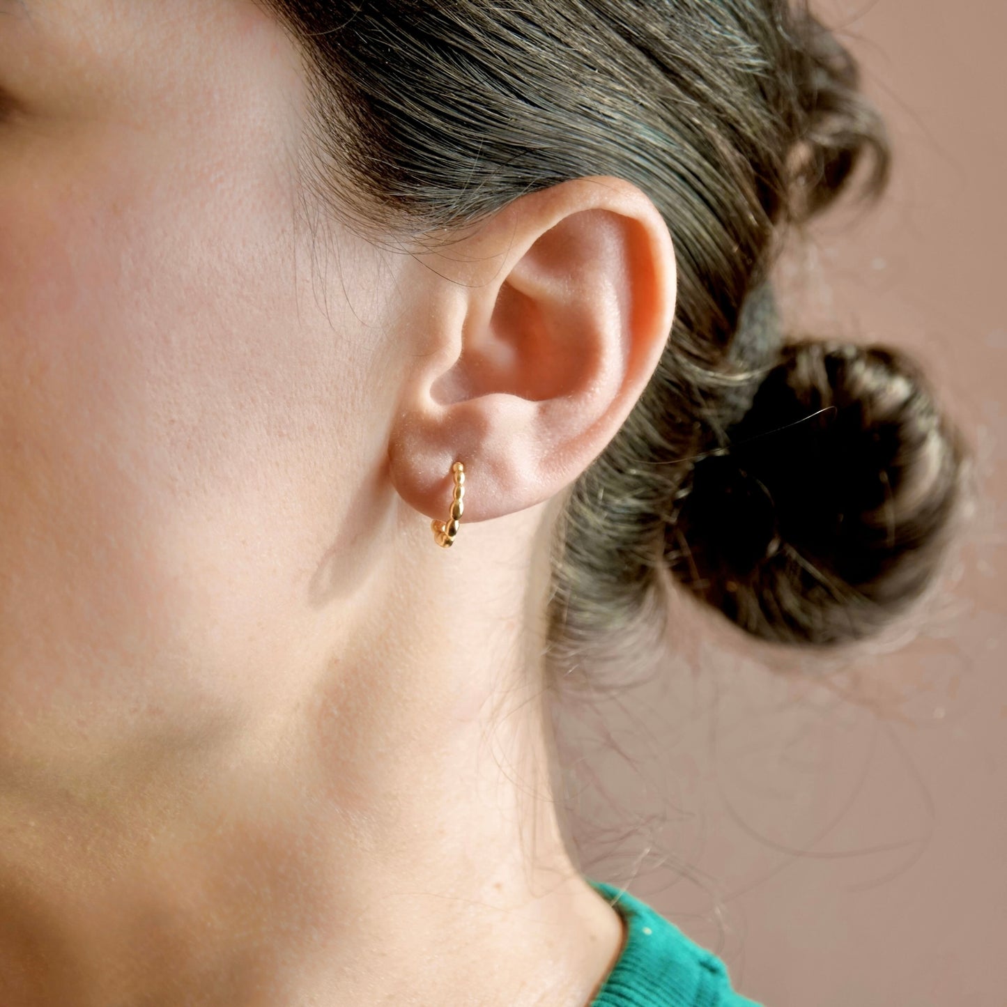 A close-up side profile of a woman wearing a small gold huggie hoop earring, with her hair in a bun against a soft warm beige background.