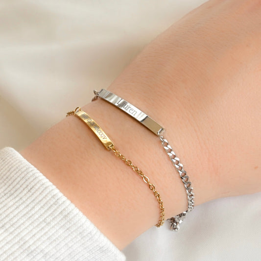 Two slim name bracelets on a person's wrist, one in gold and one in silver, with a white ribbed top visible at the wrist.