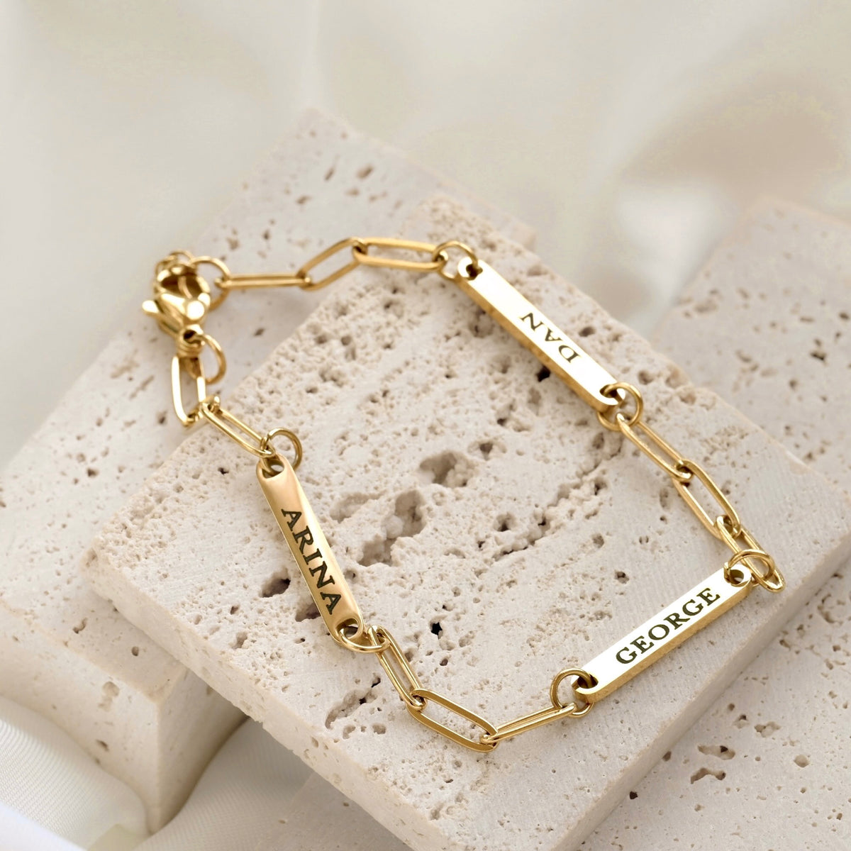 A gold paperclip chain bracelet with three rectangular nameplates engraved with the names 'ARINA', 'DAN', and 'GEORGE', displayed on stacked light beige stone blocks.