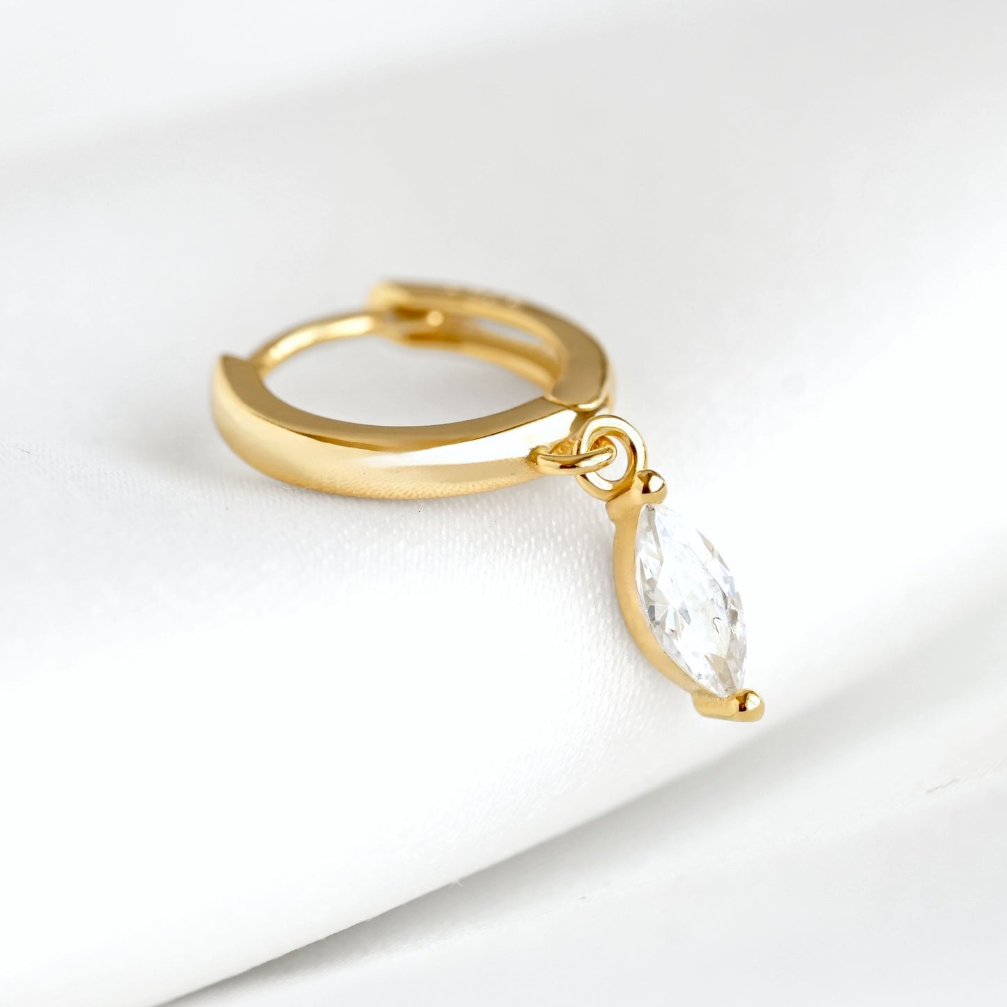 A gold huggie hoop earring with a dangling marquise-cut clear gemstone, displayed on a soft white textured surface.