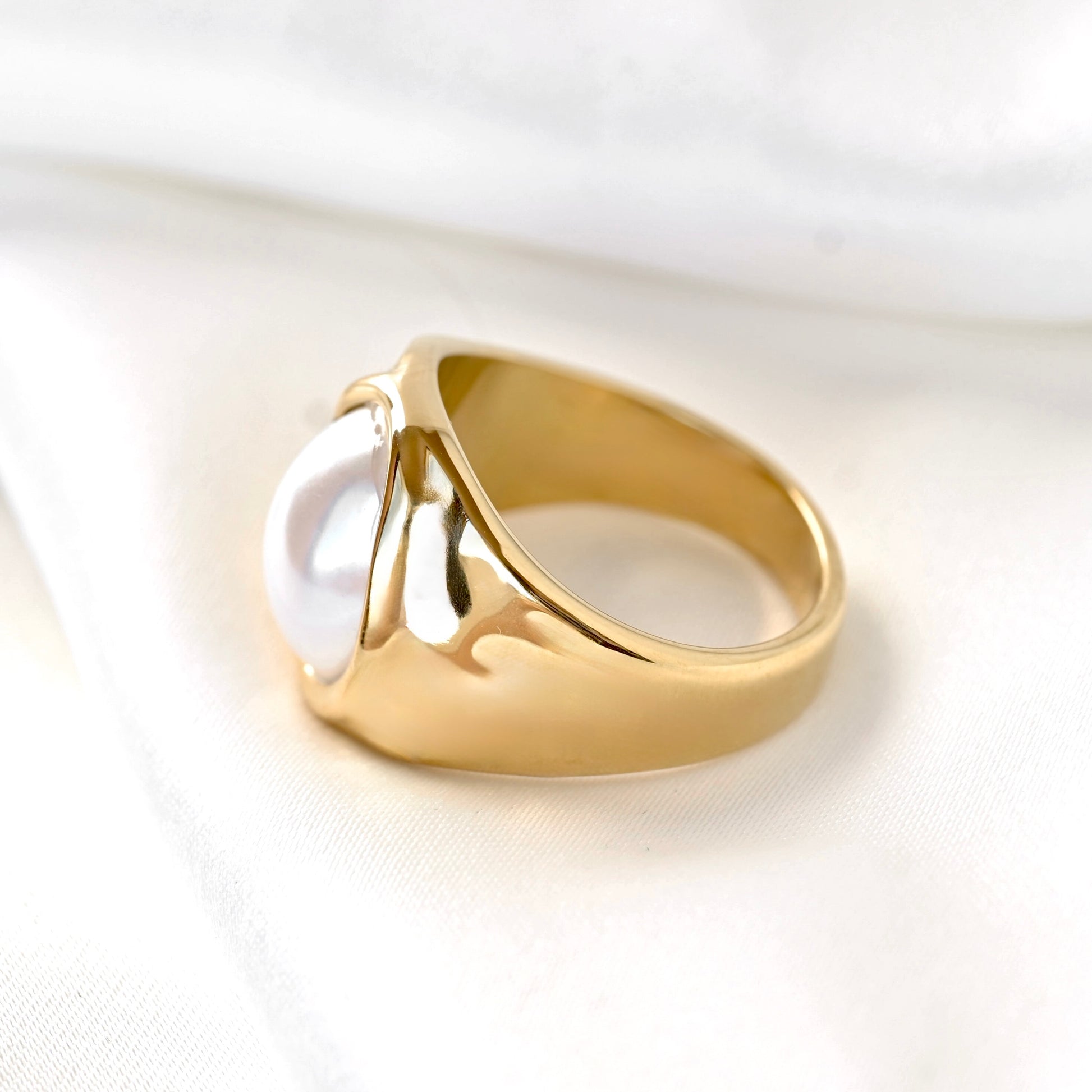 A gold ring featuring a large, smooth white pearl-like cabochon, photographed on a soft white fabric background.