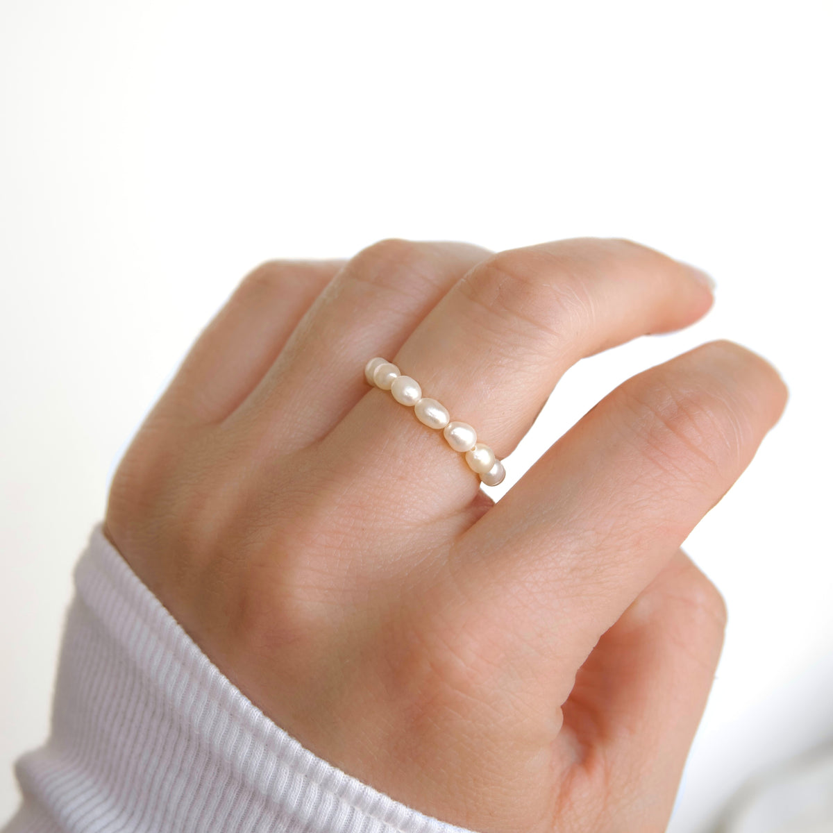A close-up of a hand wearing a delicate beaded ring made of small, round, off-white pearls. The hand is wearing a white ribbed long-sleeve top against a bright, clean white background.