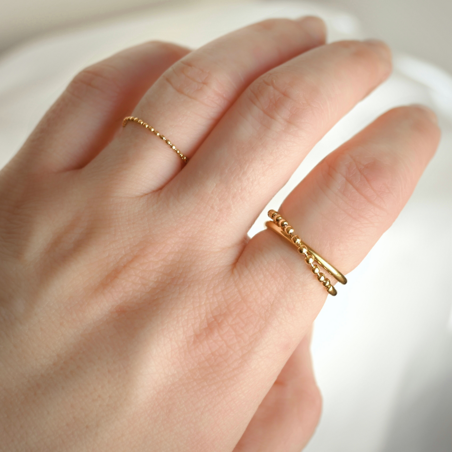 A close-up of a hand wearing two delicate gold beaded rings, with one thin chain ring on the ring finger and one textured beaded crossover band on the index finger, set against a soft white background.