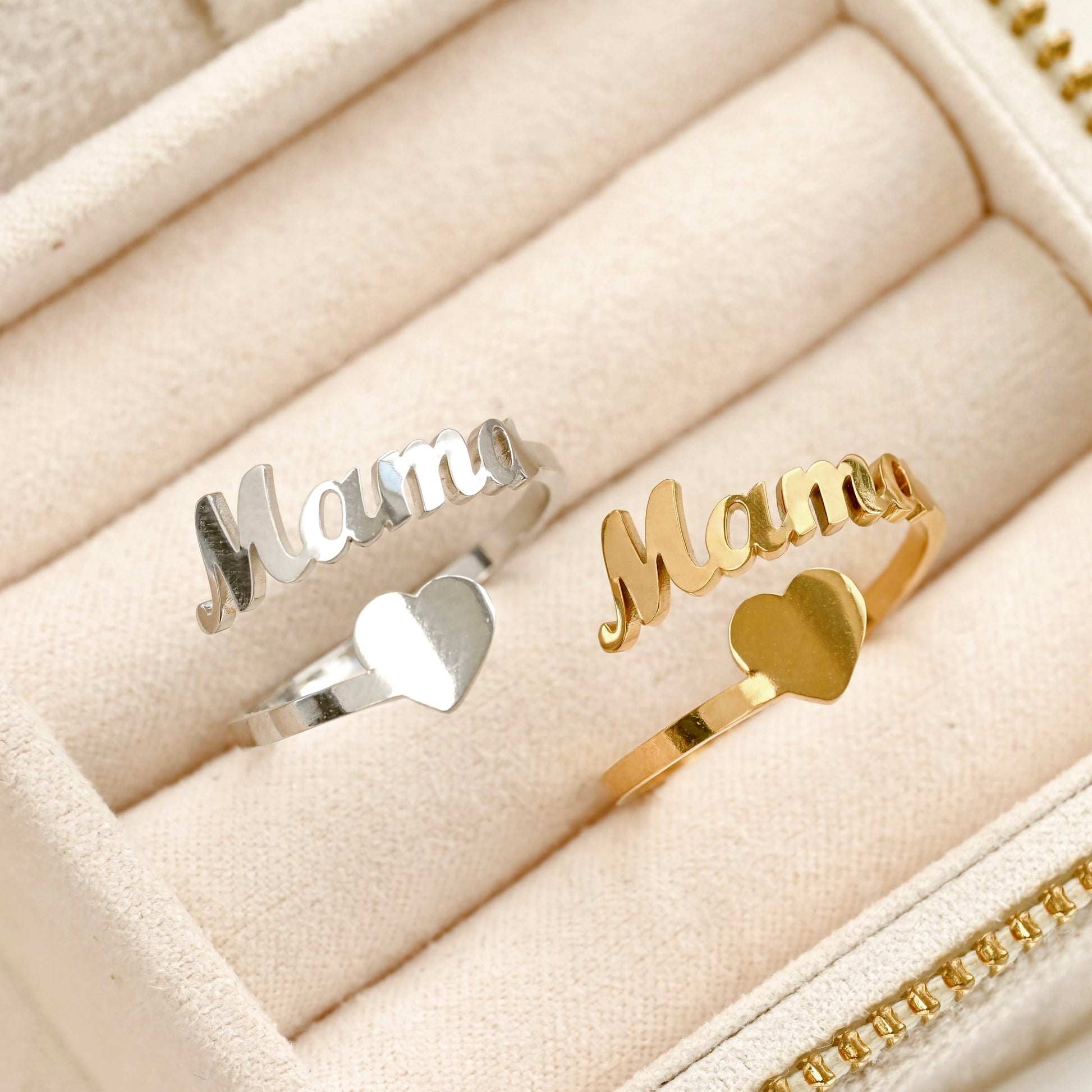 Two name rings with the word 'Mama' in cursive script and a small heart at the base, shown in silver and gold finishes inside a cream-colored jewelry box.