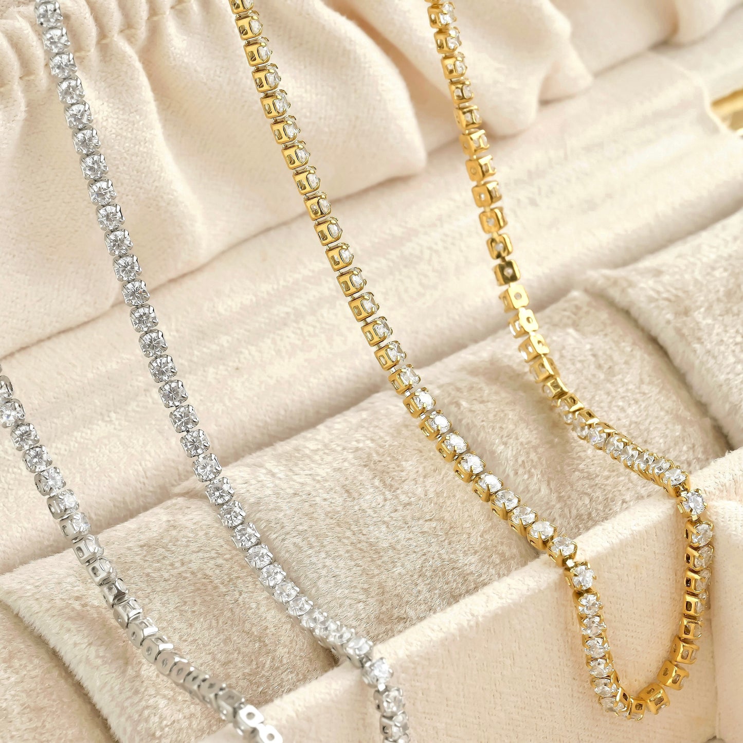 Two dainty tennis chains in gold and silver on a textured beige surface
