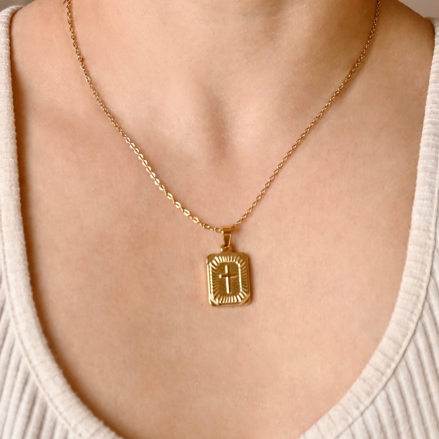A gold chain necklace with a rectangular pendant featuring a cross, worn on a model's neck over a cream-colored ribbed top.
