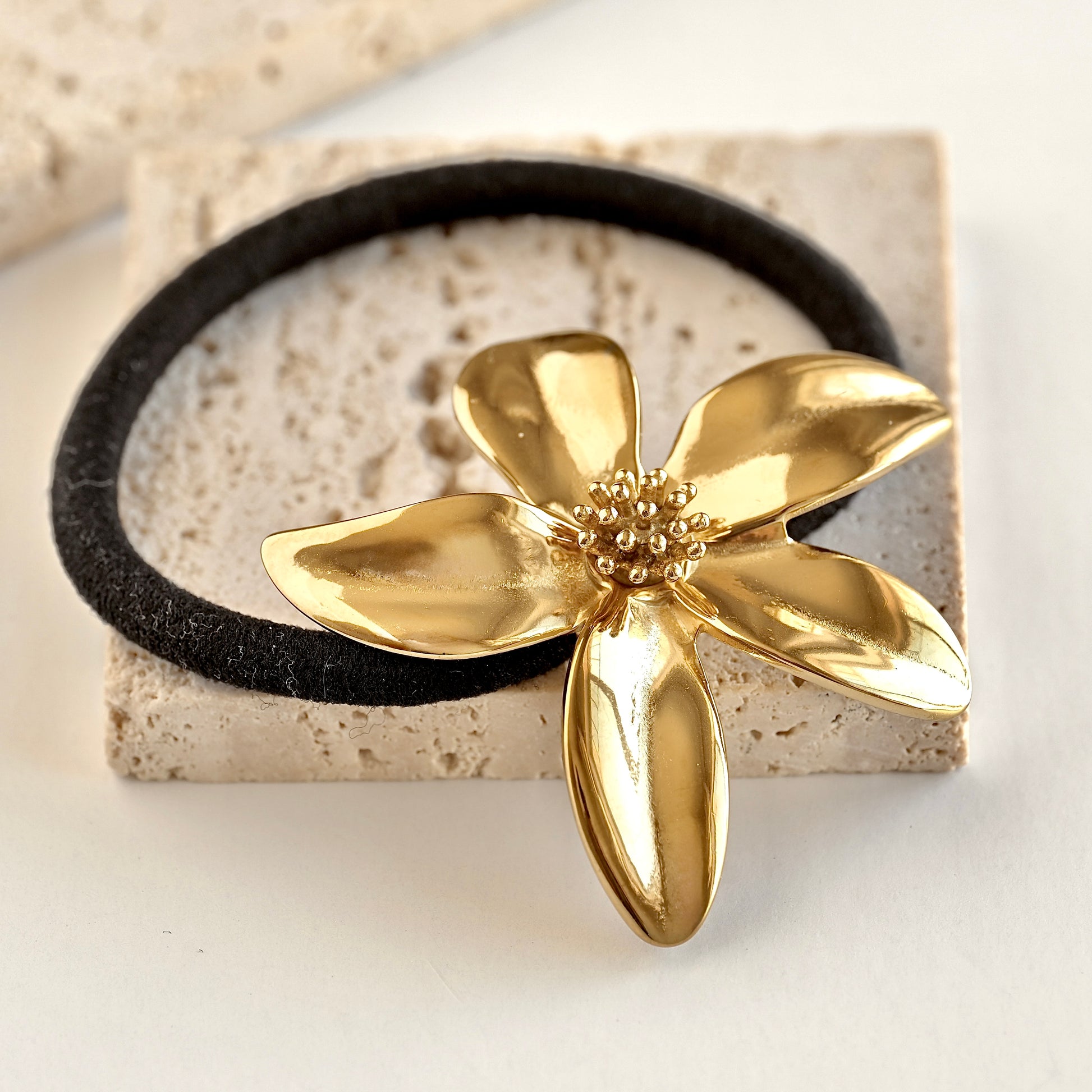 A black elastic hair tie with a large shiny gold flower-shaped hair pin attached, displayed on a beige stone block against a light neutral background.