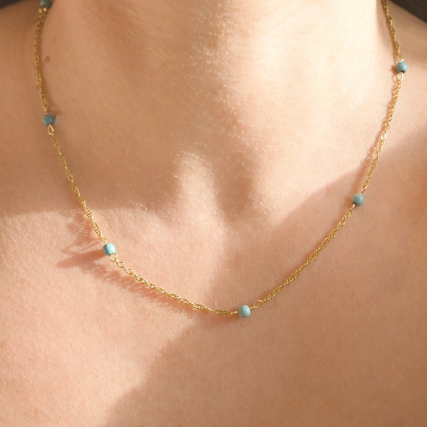A delicate gold  singapore chain necklace with small light blue beads, shown worn on a person's neck against warm sunlight.