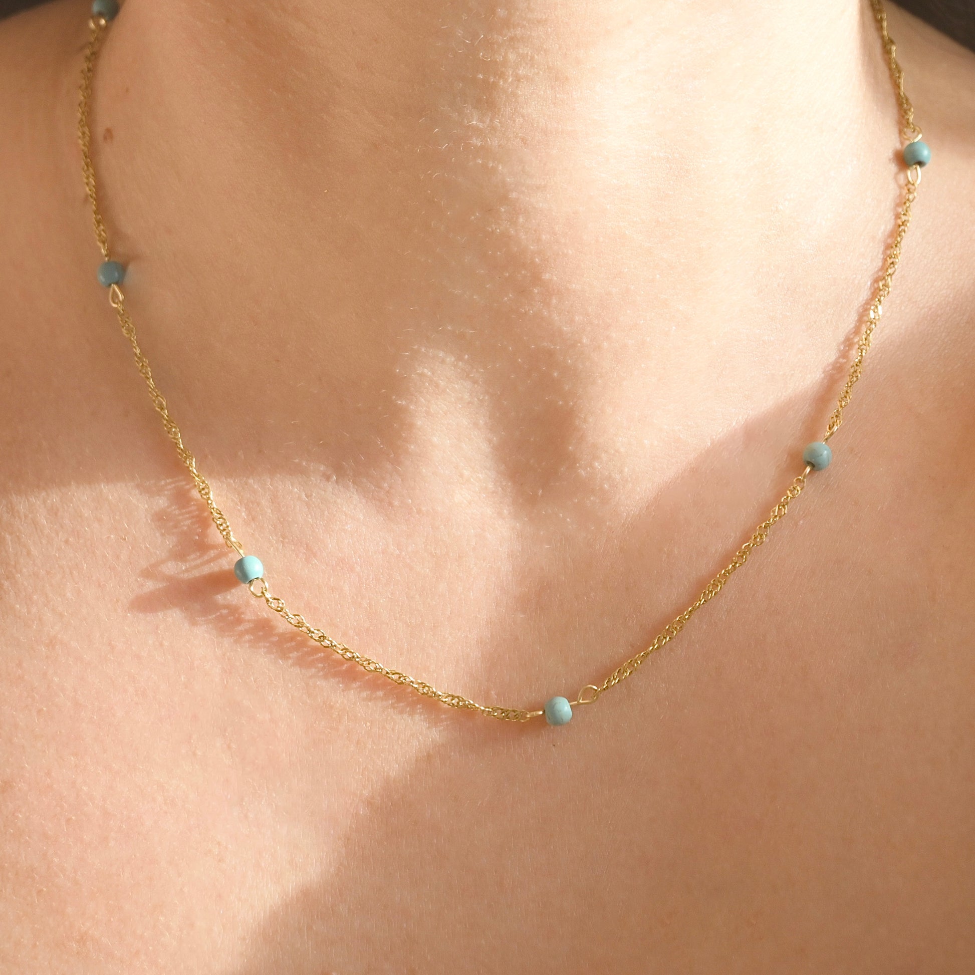 A delicate gold  singapore chain necklace with small light blue beads, shown worn on a person's neck against warm sunlight.