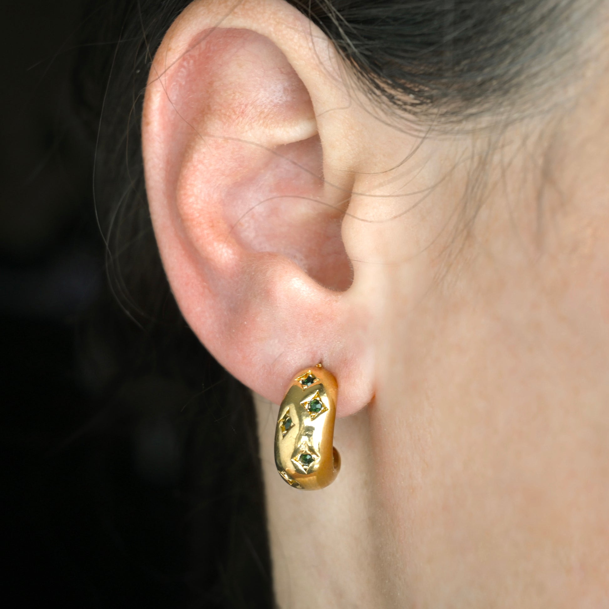 Gold hoop earring with emerald green stones worn on an ear with a dark background