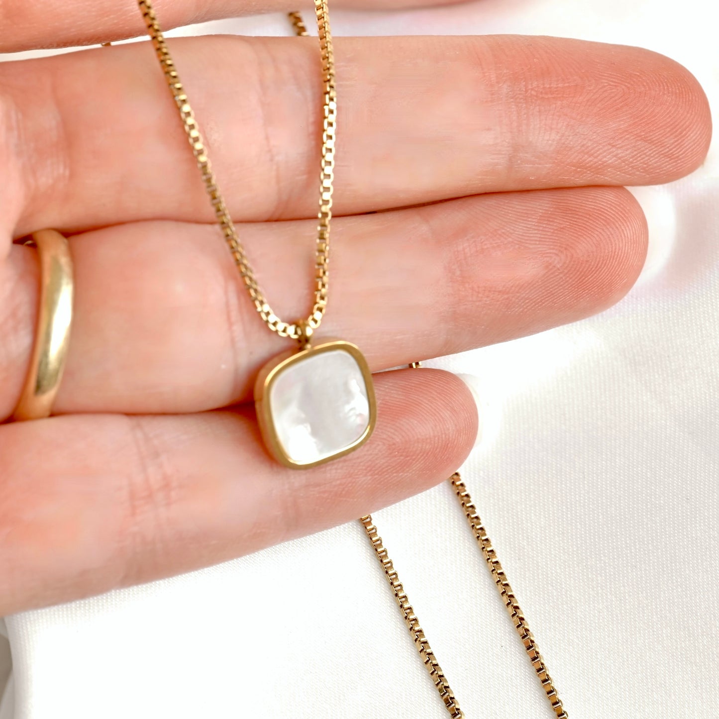 A gold chain necklace with a square pendant featuring a white mother-of-pearl inlay, held in a person's hand.