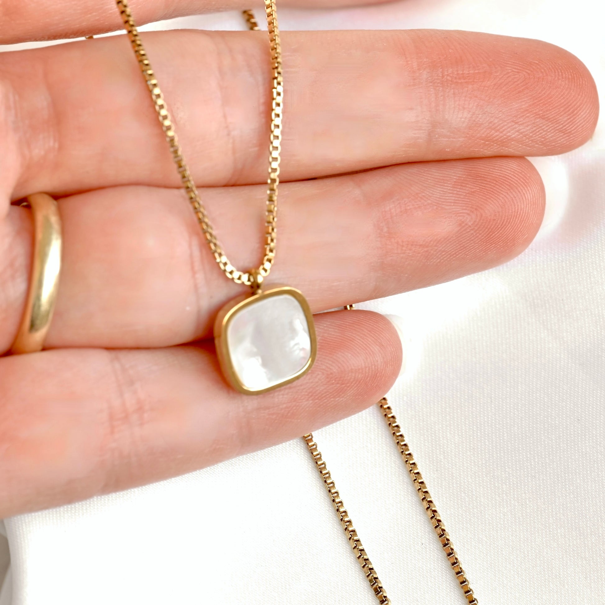 A gold chain necklace with a square pendant featuring a white mother-of-pearl inlay, held in a person's hand.